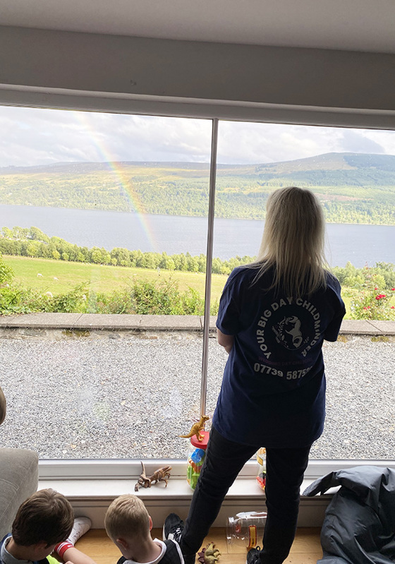 Two children play on the floor while owner of company looks out window with company logo on back of t-shirt